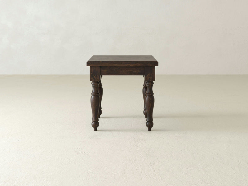 A single James+James Abigail End Table crafted from dark wood, showcasing a sophisticated turned leg motif, stands on a light beige floor against a plain, off-white wall. This rectangular table exemplifies superior craftsmanship and features detailed carvings on its legs. The setting is minimalistic and clean.