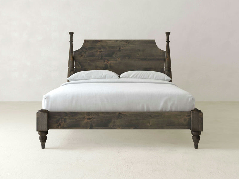 A deep grey Stella Bed from James+James featuring a tall, carved headboard and four sturdy, turned legs. The bed is neatly made with a white mattress and two white pillows, set against a plain, light-colored wall in a minimalistic room.