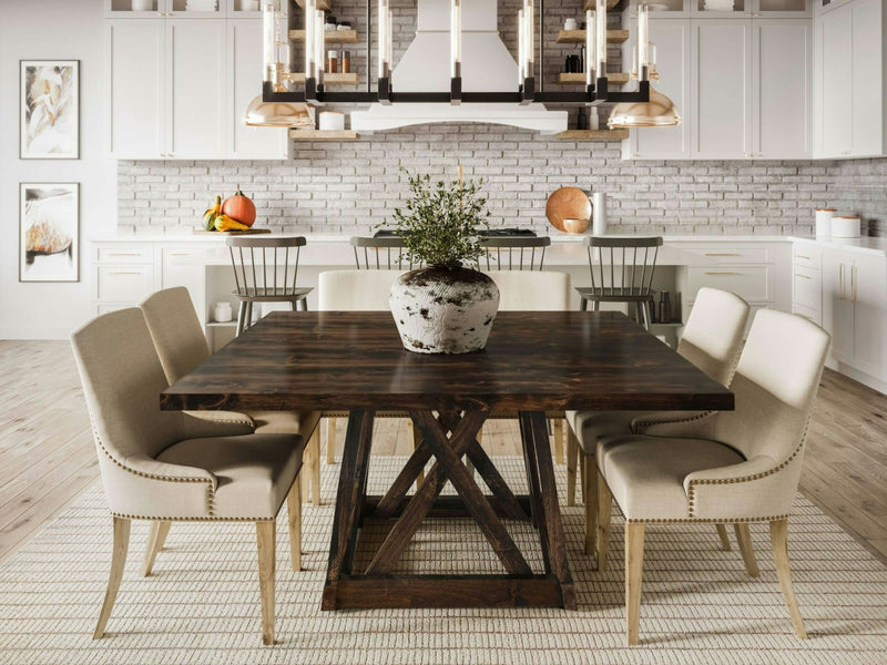 A modern dining room features the Julia Square Dining Table in Tobacco by James+James, accompanied by six beige upholstered chairs. A large plant centerpiece sits on the table. The background includes a white brick backsplash, white cabinetry, and metallic pendant lights above a kitchen island.