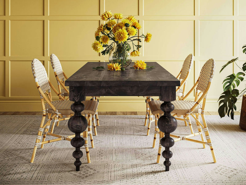 A dining room is highlighted by the Olivia Expandable Dining Table in Charred Ember from James+James, adorned with a vase of bright yellow flowers and scattered individual blooms. Surrounding the table are six wicker chairs with light-colored cushions. The room is complemented by a yellow paneled wall and a light-patterned rug.