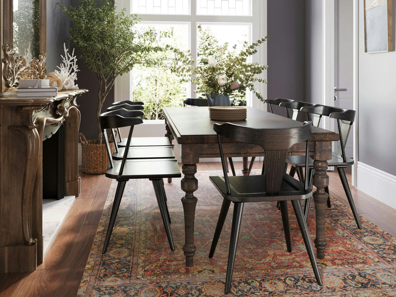 A modern dining room features the Allyson Dining Table in tobacco with five black chairs, ornate legs, and a red patterned area rug. A side console holds decor items including white coral. Large windows let in natural light, showcasing the outside greenery.