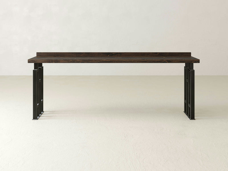 The Clark Sofa Table - Tobacco by James+James, featuring a minimalist rectangular wooden top in dark brown and supported by sleek black metal legs, stands on a plain, light-colored floor against a neutral background. The design includes a simple and clean aesthetic with a small back panel extending upward from the tabletop.