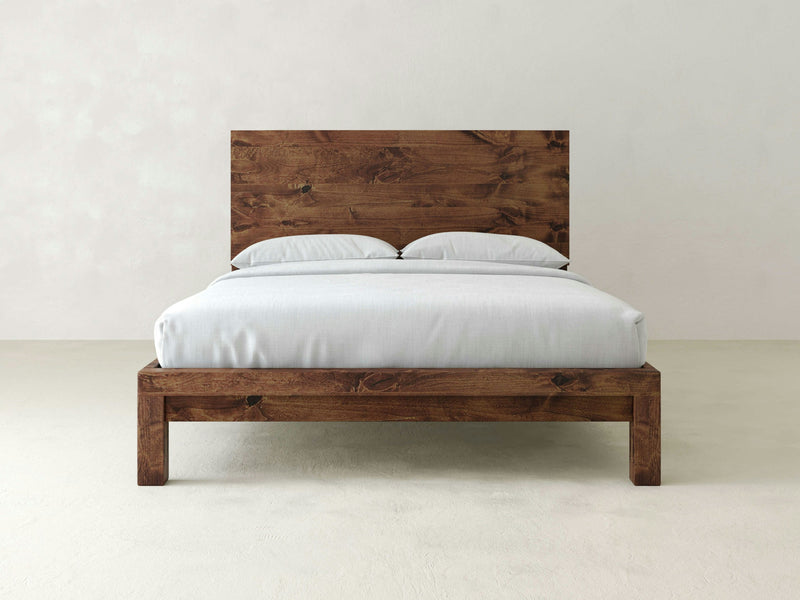 The Sawyer Bed - Tuscany by James+James, featuring a rustic wooden frame and a solid headboard, is showcased with neatly made white bedding and two pillows, all set against a plain, light-colored background.