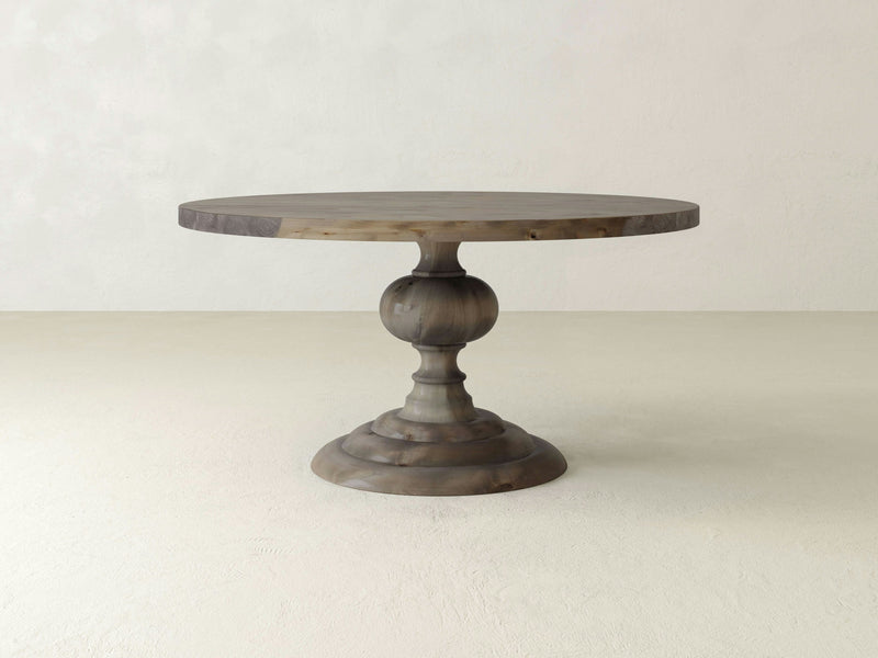 The Josephine Round Dining Table in Barn Wood by James+James stands against a plain, light-colored background. This table features a thick, sculpted pedestal base and showcases a smooth, natural wood finish with subtle grain patterns.