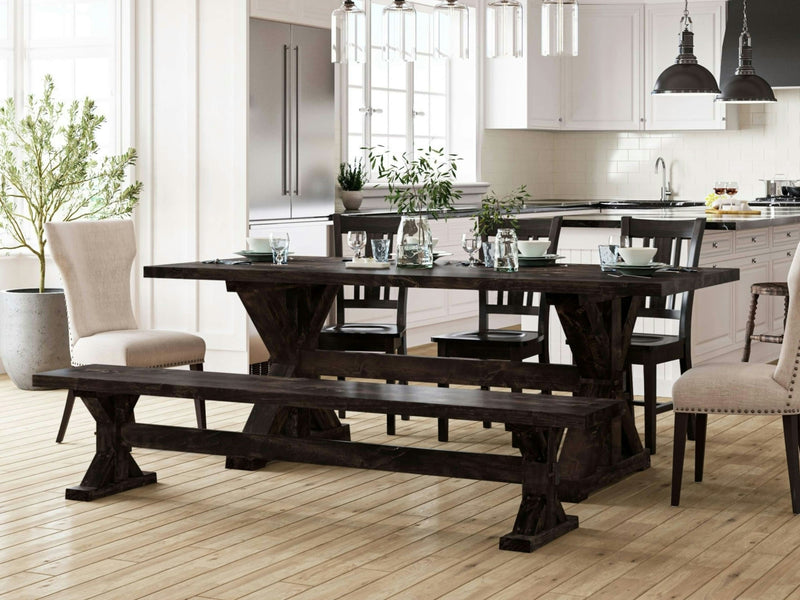 A contemporary dining area showcasing a dark wooden table with the Trestle Bench in Charred Ember by James+James, complemented by black wooden chairs. Beige cushioned chairs are situated at both ends of the table. The backdrop features a luminous white kitchen adorned with plants and pendant lights overhead.