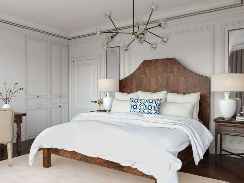 A cozy bedroom features the Megan Bed - Tuscany by James+James, adorned with white bedding and a geometric-patterned blue and white throw pillow. Two bedside tables with matching lamps flank the bed. A unique chandelier with exposed bulbs hangs from the ceiling, and a desk sits in the corner.