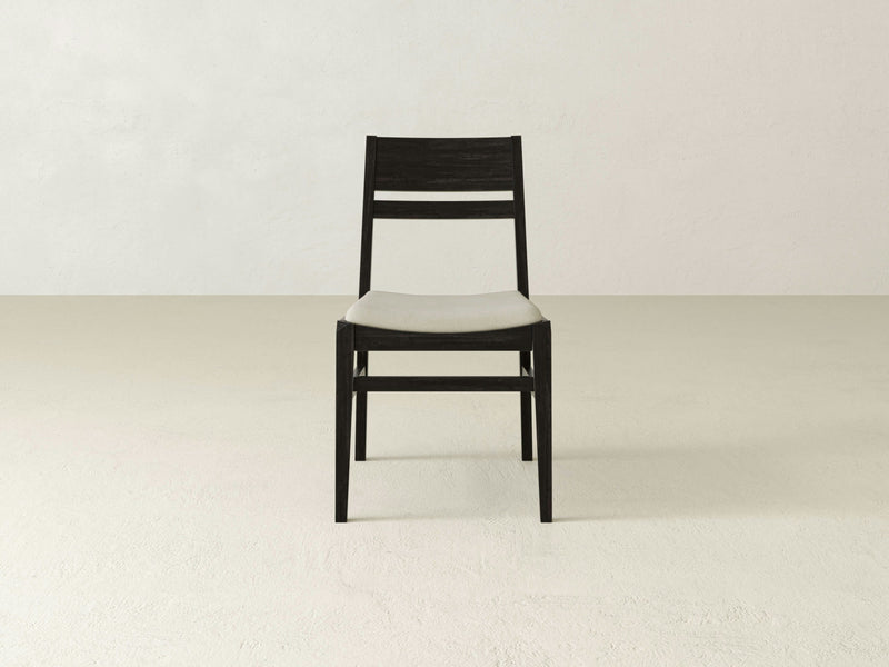 The James+James Delilah Dining Chair in Charred Ember, a minimalist black wooden chair with a light grey cushioned seat, stands on a plain, light-colored floor in front of an off-white wall. The chair's simple design features a horizontal slat backrest and straight legs.