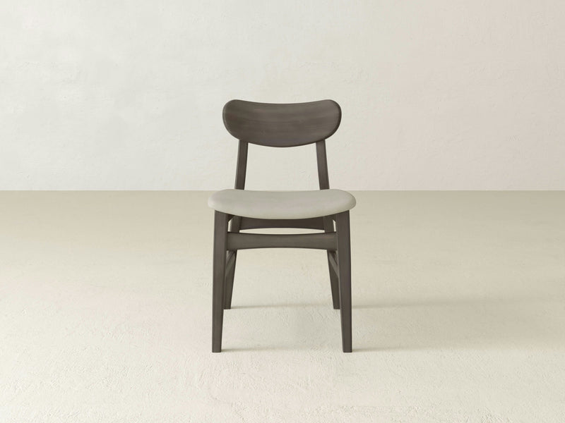 The Lynn Upholstered Dining Chair in Deep Grey by James+James features a modern wooden design with a curved backrest and a padded, light-colored seat. It is placed on a light, minimalist floor against a plain, off-white wall. The chair's simple and elegant design stands out beautifully.