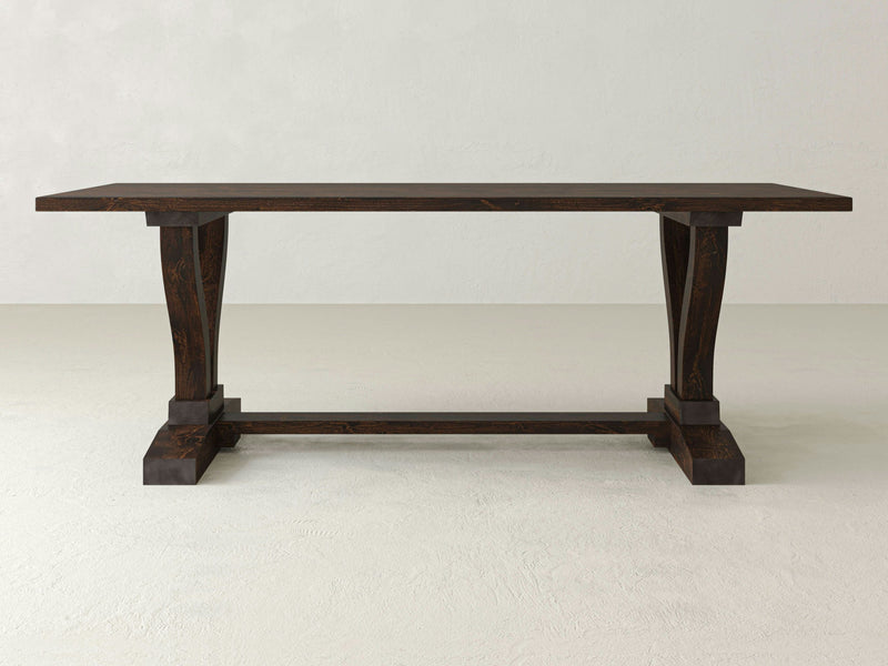 The Vera Dining Table - Tobacco by James+James features a dark wooden rectangular top and sturdy, decorative legs. The intricately carved legs are joined by a horizontal support beam. Complimented by a light, neutral background, the table's rich color and craftsmanship truly stand out.