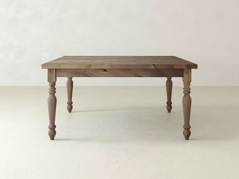 A French Country Square Dining Table - Barn Wood from James+James stands in an empty room with light-colored walls and floor. The table is crafted from medium-toned wood and boasts a simple, rustic design with a square top and four turned legs.