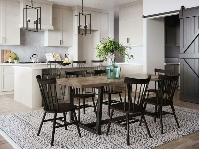 A modern dining room features the Trapezoid Dining Table in Barn Wood by James+James, complemented by six black chairs on a patterned rug. A vase with green plants sits elegantly on the table. The kitchen in the background showcases white cabinets, a light countertop, and pendant lights. A sliding barn door graces the right side of the room.