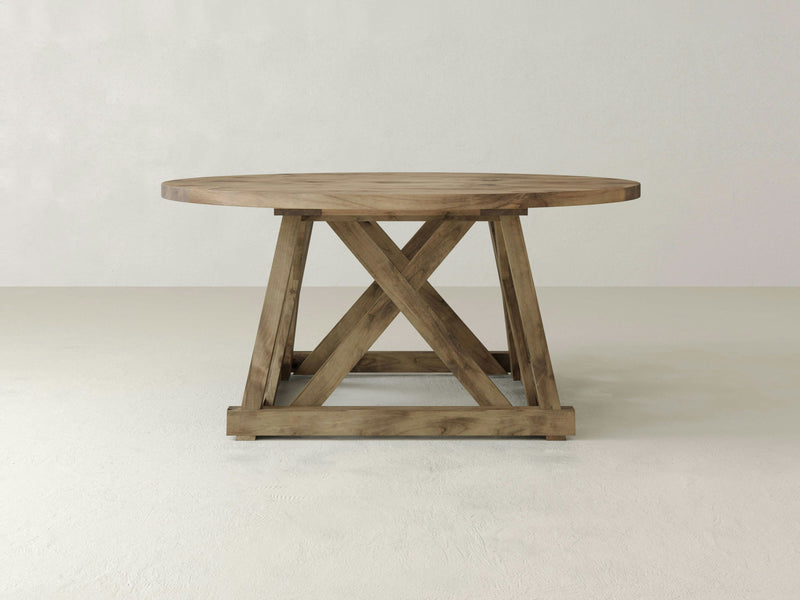 The Julia Round Dining Table by James+James, expertly crafted from solid hardwood with a rustic finish, is set against a plain, light-colored background. Its sturdy cross-brace design exudes rustic elegance and offers a robust and timeless appearance.