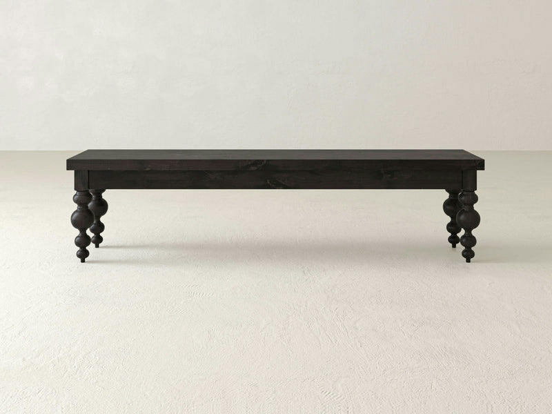 The Olivia Bench - Charred Ember by James+James features a rectangular seat and four intricately carved, spindle-style legs. The bench, finished in a smooth and polished black, boasts a simple yet elegant design that is perfect for indoor use. It stands out beautifully against the plain white background.