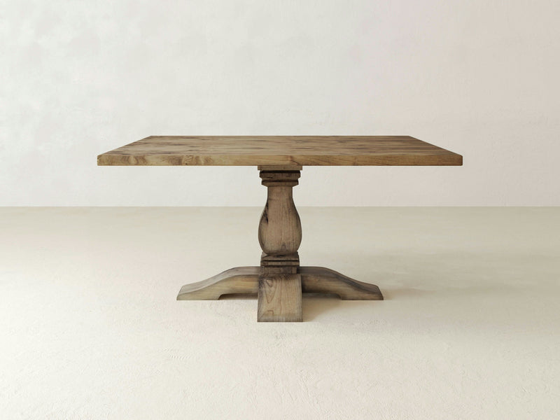 The Heirloom Square Dining Table - Harvest Wheat by James+James features a rustic wooden design with a square top and a sturdy, carved pedestal base. Set against a plain, light-colored background, the table beautifully highlights its natural wood grains and minimalist aesthetic.