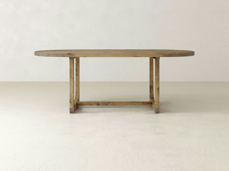 The Emersen Oval Dining Table in Harvest Wheat by James+James is centered in an empty room with a minimalist design. This oval-shaped wooden table features a sturdy double pedestal base with clean lines and exposed wood grain, placed on a smooth, light-colored floor against a plain wall.