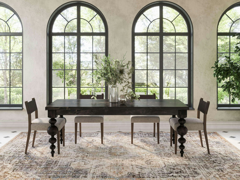 The Olivia Dining Table in Charred Ember by James+James, featuring ornate legs, is positioned in front of three large arched windows that allow ample natural light to flood the room. The table is adorned with a large floral centerpiece and surrounded by six cushioned wooden chairs. Beneath it lies a patterned rug, adding a touch of elegance to the space.