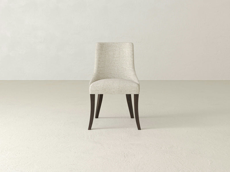 The Willow Dining Chair - Tobacco by James+James is a modern upholstered piece featuring a light beige fabric seat and dark wooden legs. Positioned against a plain light gray background, this chair boasts a gently curved backrest and slightly flared legs, exuding contemporary elegance.