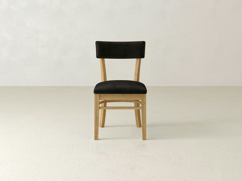 An Everly Side Dining Chair - Harvest Wheat by James+James, featuring a black cushioned seat and backrest, is placed in the center of an empty, light-colored room. The design is simple and minimalistic, showcasing light wood legs and frame. The background features a plain neutral wall and floor.