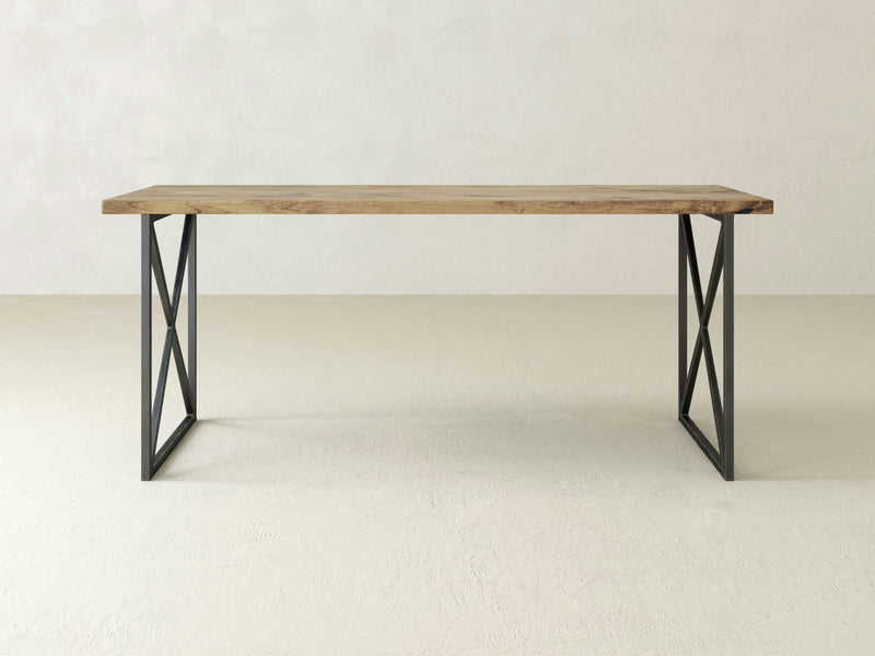 The Luca Desk - Harvest Wheat by James+James is a minimalist wooden table featuring a rectangular top and black metal legs configured in a trestle style. The distinct "X" shape on either side complements the table, which stands elegantly on a light-colored floor against a plain white wall.