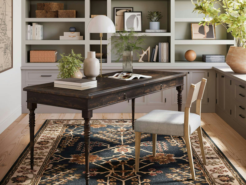 A home office with an Audrey Desk in Tobacco by James+James paired with a light grey upholstered chair. The desk has a lamp, potted plants, and books on top. The background features built-in shelves adorned with decor items and storage boxes. A decorative rug covers the wooden floor.