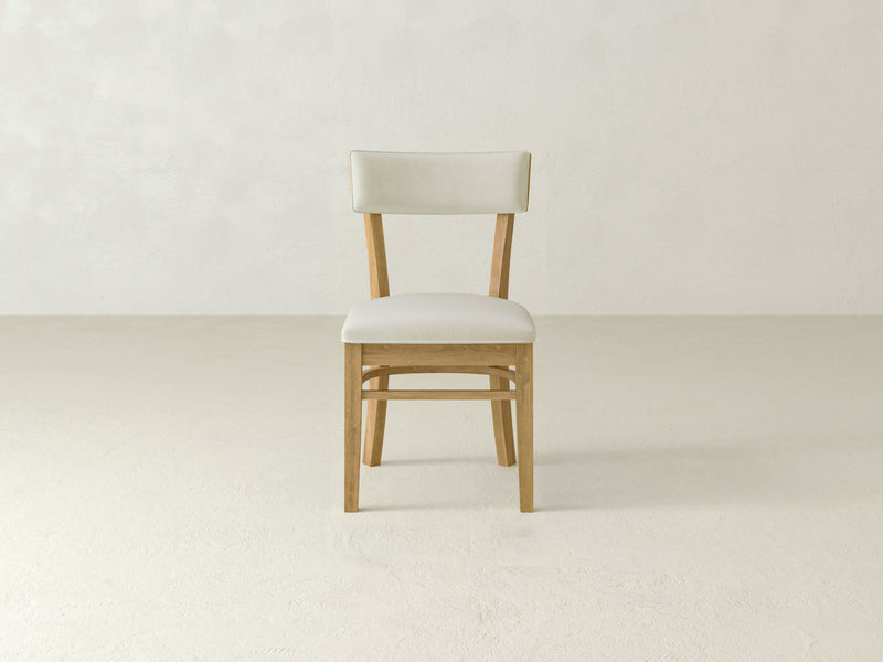 An Everly Side Dining Chair - Harvest Wheat by James+James, featuring a natural wood finish and light beige upholstered seat and backrest, is placed in the center of an empty room with a light-colored floor and wall. The design is minimalist with clean lines.