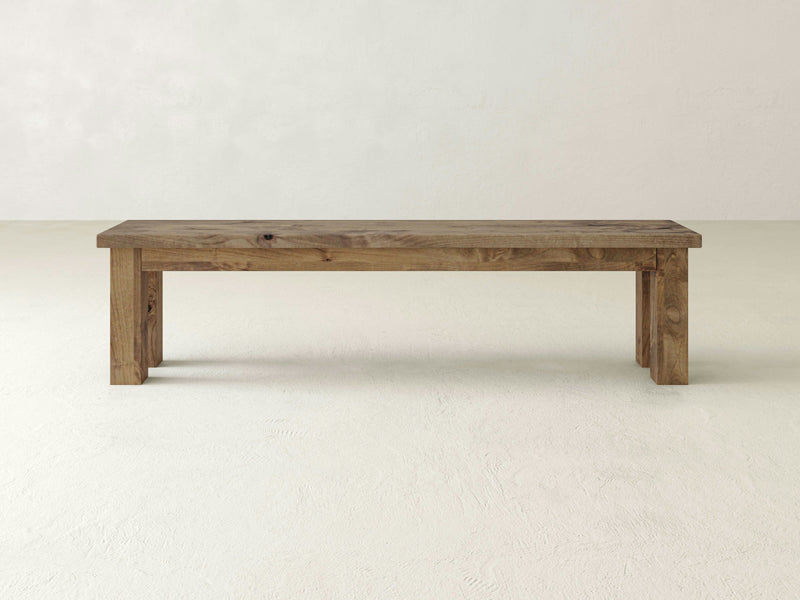 A minimalistic James+James Farmhouse Bench stands against a plain, light-colored background. Handcrafted from solid hardwood, the bench features a rectangular design with simple, sturdy legs and a natural wood grain pattern. Its clean lines emphasize functionality and rustic charm.