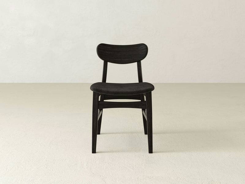 A sleek, modern Lynn Upholstered Dining Chair in Charred Ember by James+James stands in the center of a minimalist, light-colored room with a plain, off-white background.