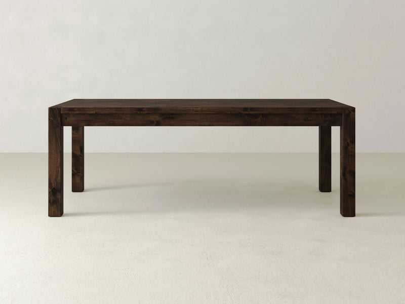 The Jonathan Dining Table - Tobacco by James+James, a minimalist dark wooden table with clean lines and a rectangular shape, stands against a plain off-white background. The table features sturdy legs and a smooth, polished surface, showcasing a simple yet elegant design suitable for various interior settings.