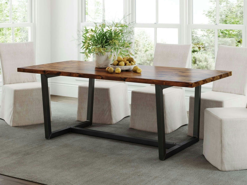 A Trapezoid Dining Table - Tuscany from James+James, featuring a black metal base and a wooden top, is centered in a room with large windows. The table is decorated with a white vase filled with green plants and a bowl of lemons. Four cushioned beige dining chairs surround the table, which sits on a gray carpet.