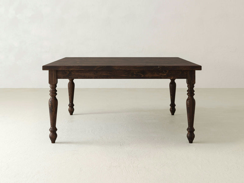 A French Country Square Dining Table in Tobacco finish from James+James stands on a light-colored floor against a plain, light-colored wall. The table features ornate, carved legs and a square top with a classic design, giving it a vintage or rustic appearance.