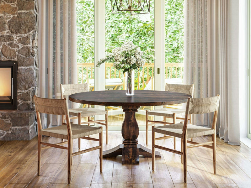 The Heirloom Round Dining Table - Tobacco by James+James is a stunning piece with a dark brown finish. It boasts a thick, solid tabletop and an ornate single pedestal base, featuring four carved legs that extend outward for support. The design is both rustic and elegant, placed against a plain white background.