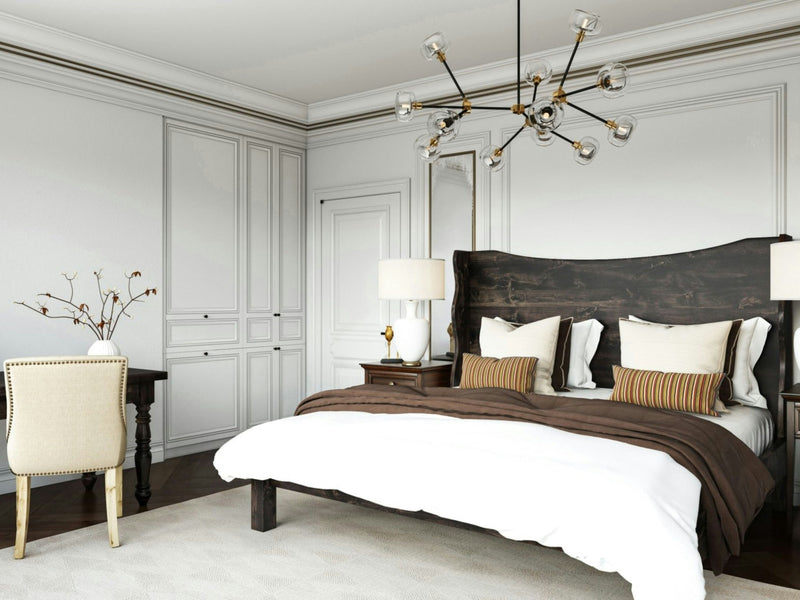 A modern bedroom with a James+James Harlow Bed in Charred Ember, adorned with beige bedding on a double bed. The room features a white door, built-in white cabinets, and a stylish chandelier. A beige chair sits by a dark wooden desk with a vase holding branches. Soft lighting fills the room.