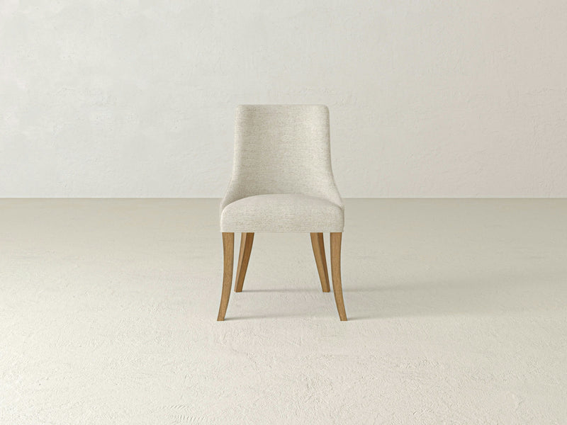 A modern Willow Dining Chair - Harvest Wheat by James+James, featuring a light beige color and minimalist design with wooden legs, is centered against a plain, light-colored background. The chair boasts a padded seat and backrest with gently curved lines and a simple, elegant silhouette.