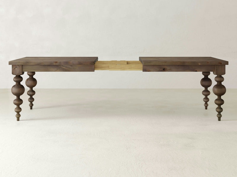 Introducing the Olivia Expandable Dining Table - Barn Wood by James+James, featuring intricately carved, baluster-style legs. Shown in its extended state, the table's middle section highlights a striking lighter wood contrast. The background provides a clean look with its plain, neutral-colored wall and floor.