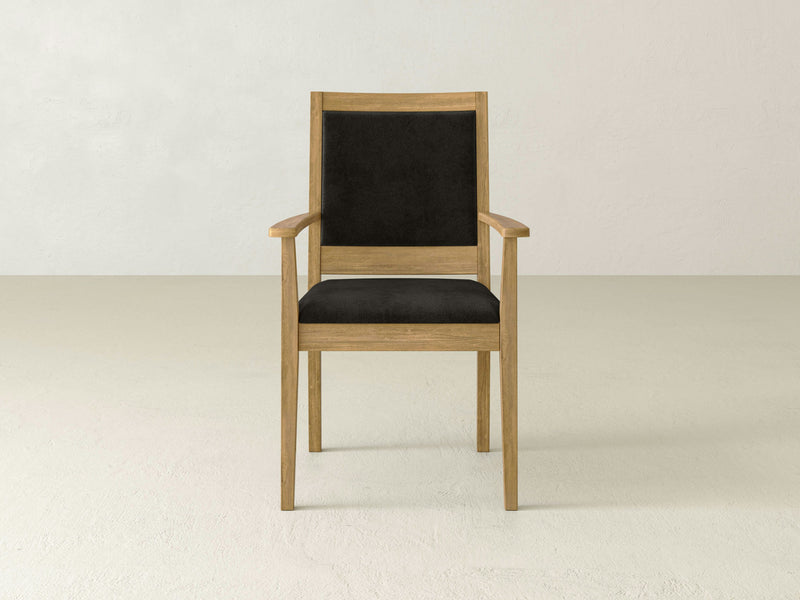 The Gina Arm Dining Chair - Harvest Wheat by James+James, featuring a minimalist wooden frame with black upholstered seat and backrest, is centrally placed against a plain light-colored wall and floor background. The chair boasts a simple yet sturdy design with armrests.