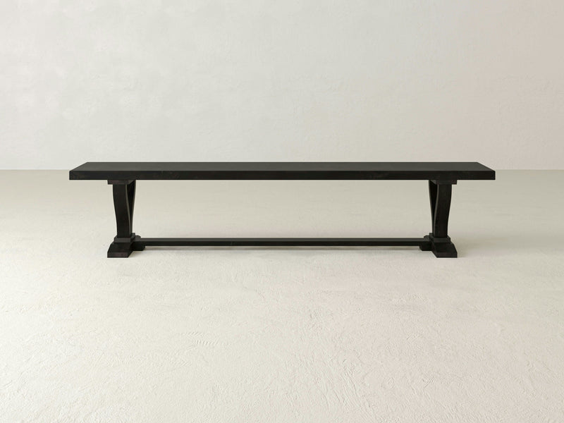 A minimalist Vera Bench - Charred Ember by James+James stands alone in a white, spacious room. This dark wooden bench with clean lines features simple, rectangular legs and a supportive bar connecting the legs at the base, creating a sturdy yet elegant piece of furniture.