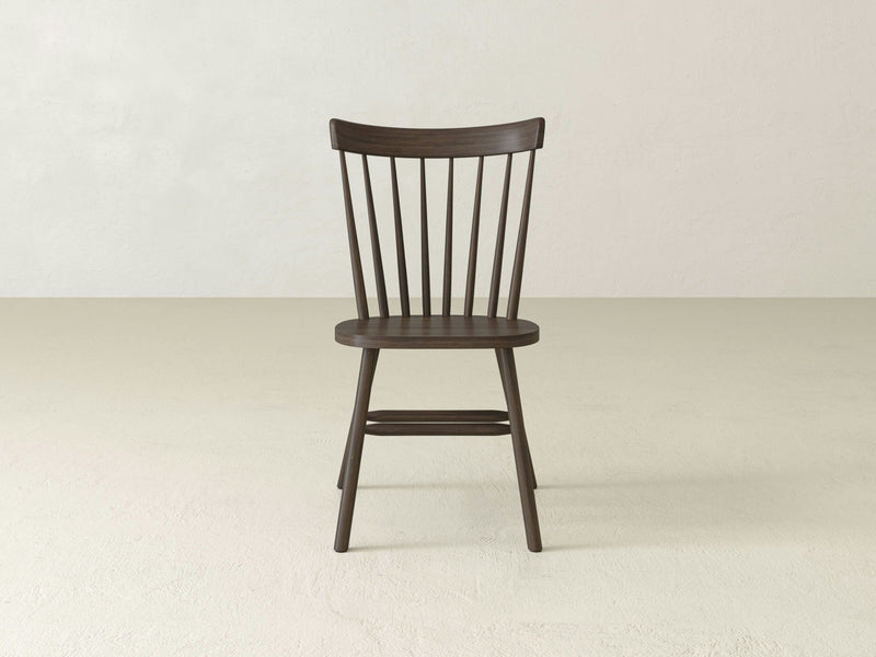 The Rustic Windsor Dining Chair - Tobacco by James+James is centered in the image. This wooden chair features a simple design with a slightly curved backrest and vertical slats, finished in dark brown. The seat is smooth, and the tapered legs are connected by horizontal supports. The background is a light, plain wall and floor.