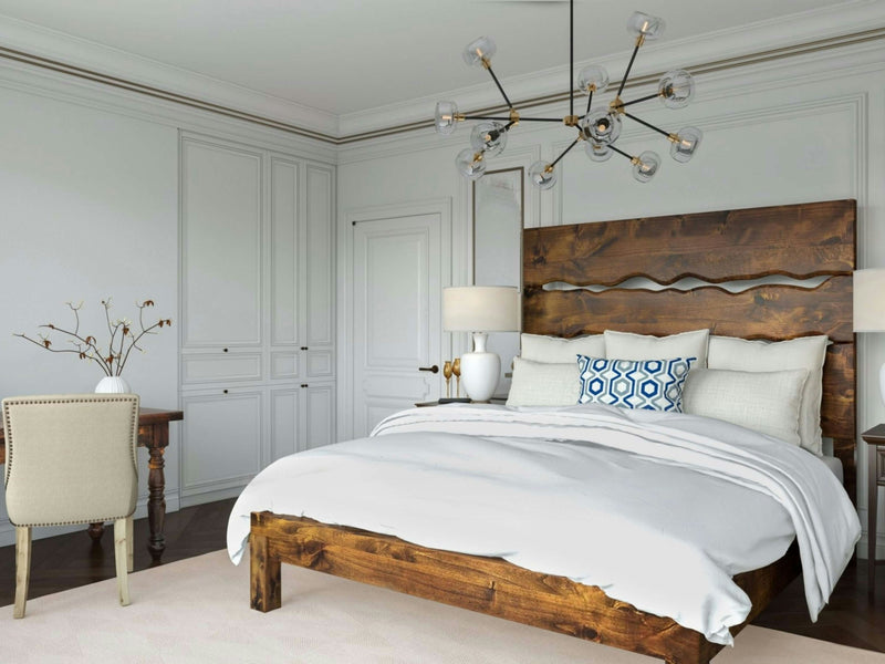 A modern bedroom showcases the Live Edge Bed - Tuscany by James+James, adorned with white bedding and a blue patterned pillow. Hanging above the bed is a contemporary chandelier. A desk with a beige chair is situated to the left, while the room's walls are painted light grey and feature built-in closets.