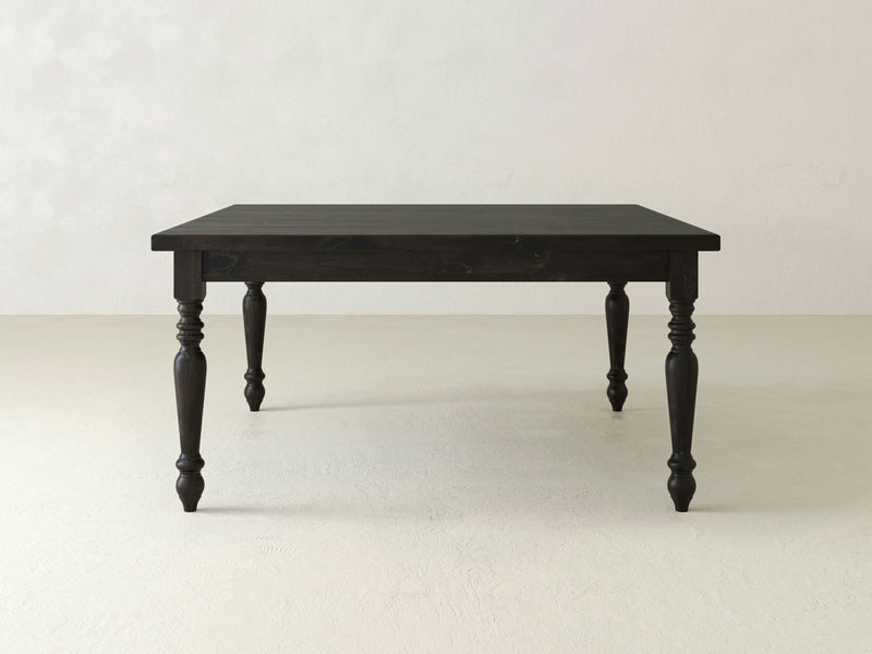 The French Country Square Dining Table in Charred Ember from James+James, featuring a dark wooden rectangular top and four elegantly turned legs, is set against a plain, light-colored background. This table boasts a simple and classic rustic design.