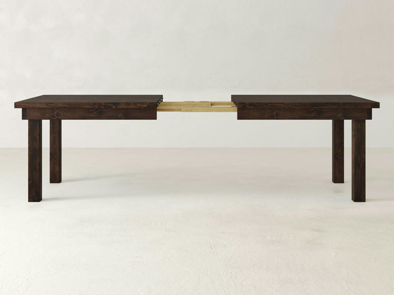 The Farmhouse Expandable Dining Table - Tobacco by James+James is shown in an expanded state, revealing its extension mechanism in the middle. The table features four sturdy legs and a simple, minimalist design. The plain, light-colored background enhances the dark wood's details and highlights the table's features.