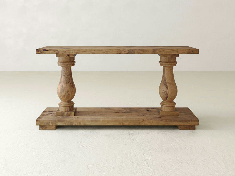 The James+James Vivien Sofa Table in Harvest Wheat features a rectangular top and bottom shelf, supported by two ornately carved, wide cylindrical legs. Its natural wood finish with visible grain patterns beautifully combines traditional and rustic elements.