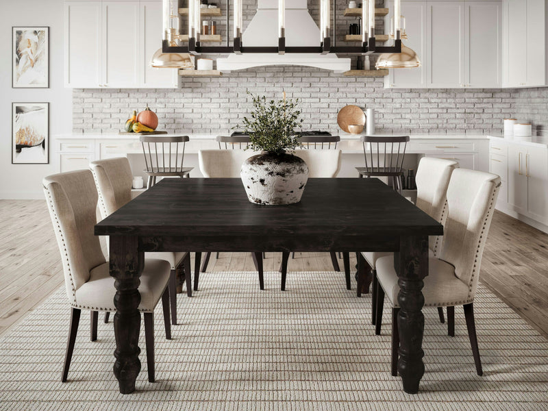 A modern dining room featuring a James+James Baluster Square Dining Table in Charred Ember, accompanied by eight beige upholstered chairs. The table is adorned with a large potted plant in its center. The background showcases white cabinetry, a white brick backsplash, pendant lights, and decorative elements such as framed art and a bowl of fruit.