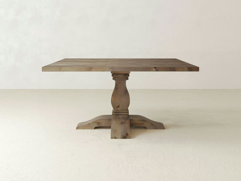 The Heirloom Square Dining Table - Barn Wood from James+James, featuring a decorative pedestal base, stands on a plain light-colored floor against a light-colored wall. The table boasts a simple, rustic design with a natural wood finish.