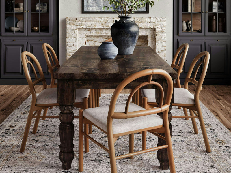 A dining room features the Baluster Dining Table - Tobacco by James+James, surrounded by six wooden chairs with light-colored cushions. The table holds two decorative vases. In the background, there's a white brick fireplace and dark cabinetry. The floor has a patterned rug.