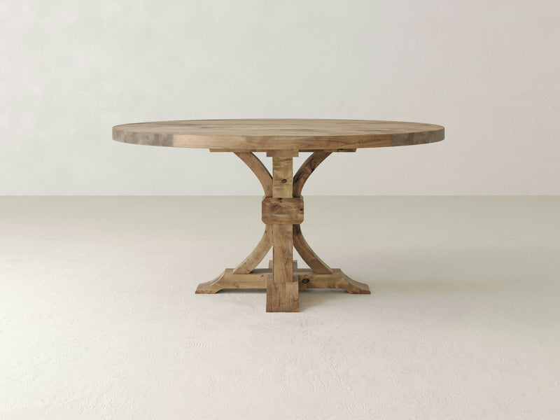 Introducing the Violet Round Dining Table - Harvest Wheat by James+James: an elegant wooden table featuring a smooth top surface and a robust four-legged pedestal base. The table showcases a natural wood finish, beautifully highlighting its grain and texture. It stands on a light-colored floor against a plain, off-white background.