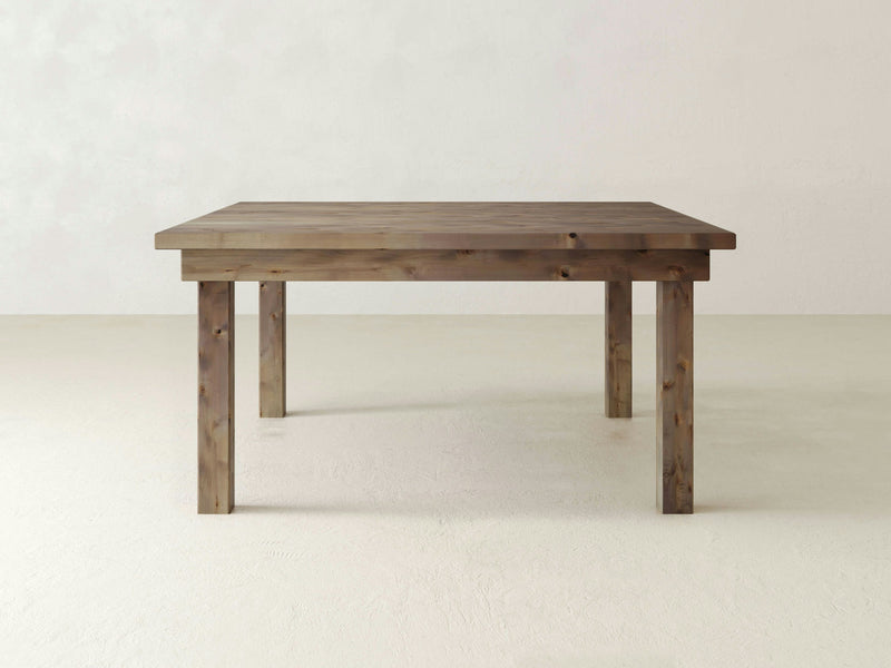 The Farmhouse Square Dining Table - Barn Wood from James+James is centered in a minimalistic room with light-colored walls and floor. This simple yet elegant wooden dining table features sturdy, rectangular legs and a plain, flat tabletop with a natural finish.