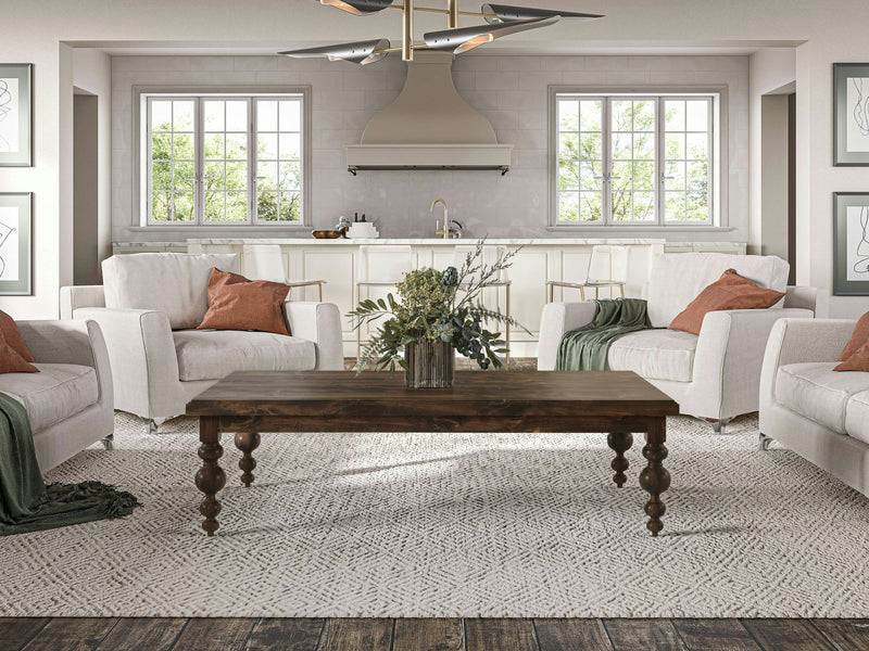 A cozy living room featuring four white armchairs adorned with green and brown throw pillows, arranged around the Olivia Coffee Table – Tobacco by James+James. The room boasts large windows, lush green plants, and a light-colored rug on a wooden floor. In the background, a kitchen with two windows is visible.