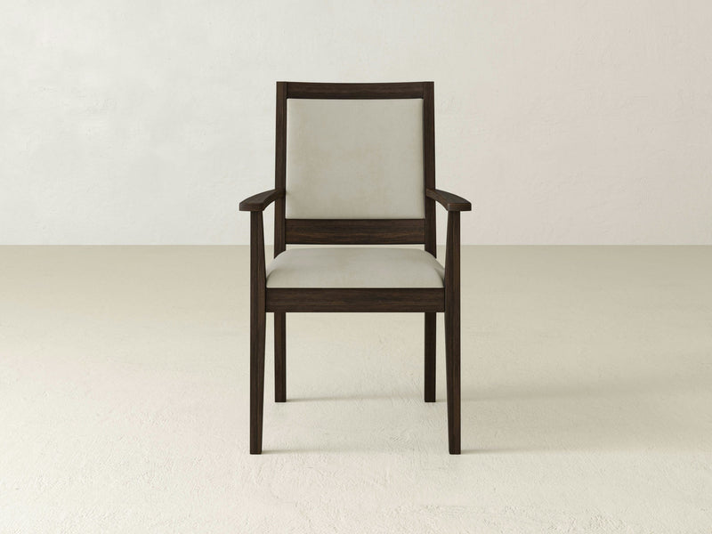 The Gina Arm Dining Chair - Tobacco by James+James, featuring a dark brown frame and light beige upholstery on the seat and backrest, is centered in a minimalist setting with off-white textured walls and a plain light floor. The design is simple and contemporary with clean lines.