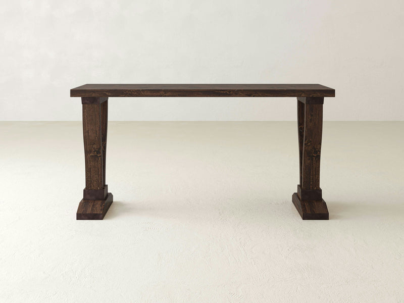 A Vera Sofa Table - Tobacco by James+James, featuring dark wood with thick, carved legs and a minimalist design, stands against a plain off-white background. The table exudes a rustic appearance and is shown from the front, emphasizing its sturdy construction and simple elegance.