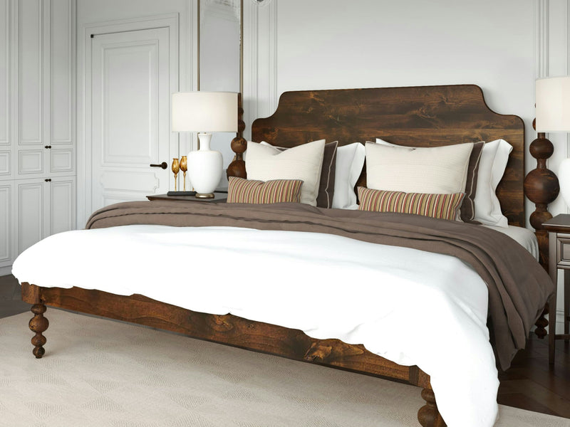 A bedroom featuring the Opal Bed in Tobacco from James+James, showcasing an intricately carved wooden headboard. The bed is adorned with white pillows, two striped accent pillows, a white duvet, and a brown throw blanket. Two matching bedside tables hold white lamps. The walls are painted white.